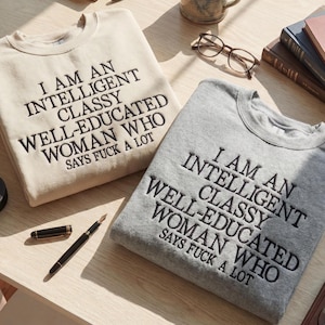 May include: Two crewneck sweatshirts, one beige and one gray, embroidered with the phrase "I AM AN INTELLIGENT CLASSY WELL-EDUCATED WOMAN WHO SAYS FUCK A LOT" in black thread. A fountain pen and glasses are also visible.