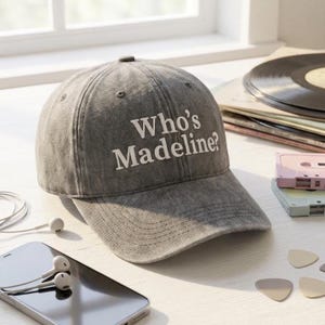 May include: Gray baseball cap with the text "Who's Madeline?" in white. The cap is made of a soft, textured material. The image also includes a smartphone, earbuds, and a stack of vinyl records.