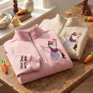May include: Two zip-up sweatshirts, one pink and one beige, with an embroidered design of Jesus Christ swinging on an Easter egg. The pink sweatshirt has the text "Jesus Christ, you're back!" on the cuff. Easter-themed apparel.
