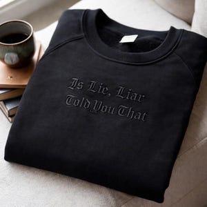 May include: Black crew neck sweatshirt with the embroidered text "Is Lie, Liar Told You That". The sweatshirt is folded on a light-colored surface, with a cup of coffee and books in the background.