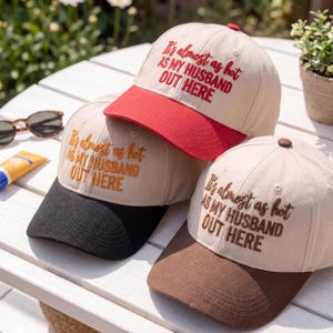 May include: Three baseball caps with the text "It's almost as hot as my husband out here." The caps have tan crowns and coloured brims in red, black, and brown. A small plant and sunglasses are in the background.