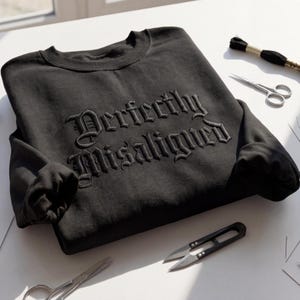 Op de afbeelding: Zwart sweatshirt met ronde hals en de geborduurde tekst "Perfectly Misaligned" in gotische letters. De afbeelding toont borduurbenodigdheden, waaronder een schaar en draad, op een wit oppervlak.