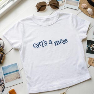 May include: White t-shirt with the embroidered text "Carl's a mess" in navy blue. The shirt is short-sleeved and has a crew neck. Sunglasses, film roll, and photos are arranged around the shirt on a white surface.