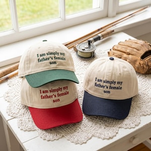 Pode incluir: Três bonés de beisebol com copa cor de creme e aba colorida. Um boné tem aba verde-escura com texto verde, um tem aba vermelha com texto vermelho e um tem aba azul-marinho com texto azul-marinho. Todos os bonés apresentam o texto bordado: I am simply my father's female son.