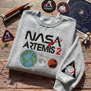 May include: A light gray sweatshirt with the NASA Artemis 2 logo embroidered in black and red. The design includes Earth, the Moon, and Mars. A patch with a rocket ship is on the sleeve. Patches and a model rocket are also visible.