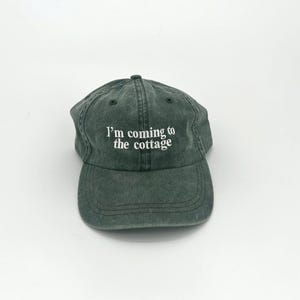 May include: A faded green baseball cap with a curved brim. The cap features the white text, "I'm coming to the cottage" embroidered on the front. The hat is made of a soft, worn material.