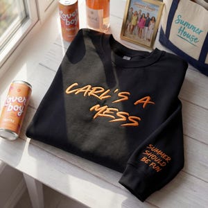 May include: Black sweatshirt with the embroidered text "CARL'S A MESS" in orange. The sleeve reads "SUMMER SHOULD BE FUN". The image also includes two cans of "loverboy spritz", a bottle of rosé, a framed photo, and a tote bag.