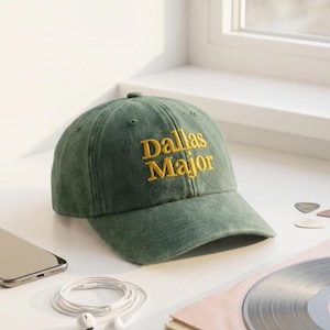May include: A green baseball cap with the text "Dallas Major" embroidered in yellow. The cap is made of a soft material and has a curved brim. The cap is sitting on a white surface next to a smartphone, earbuds, and guitar picks.