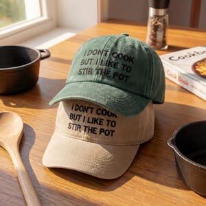 May include: Two baseball caps, one green and one tan, stacked on top of each other. The green cap has the text "I DON'T COOK BUT I LIKE TO STIR THE POT" embroidered on the front. The tan cap has the same text.