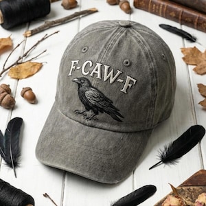 May include: A gray baseball cap with the embroidered text "F-CAW-F" and a detailed black raven illustration. The cap is made of a worn fabric and has a curved brim. The background includes black feathers and acorns.