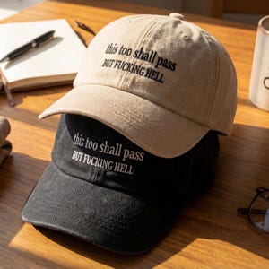 May include: Two baseball caps, one tan and one black, stacked on top of each other. Each cap has the embroidered text "this too shall pass BUT FUCKING HELL" in black and white, respectively. The caps are on a wooden surface.