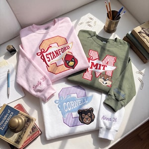 May include: Three crewneck sweatshirts in light pink, olive green, and white. Each features a large collegiate letter with a pennant and mascot embroidery. The pink sweatshirt says Stanford, the green says MIT, and the white says Cornell. Sleeves are embroidered with names and graduation years.