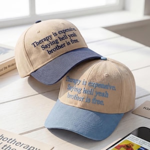 May include: Two beige baseball caps with blue brims are displayed. Each cap has the embroidered text: "Therapy is expensive. Saying hell yeah brother is free."