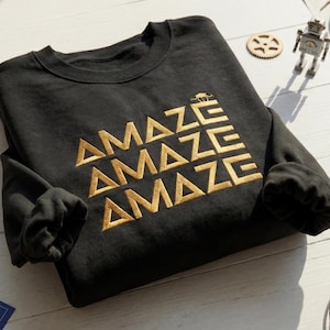 May include: Black sweatshirt with the word "AMAZE" in gold block letters repeated three times. The sweatshirt is folded, with the sleeves tucked in. A small kangaroo graphic is above the word "AMAZE".