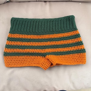 May include: Handmade crochet shorts in a vibrant color scheme. The shorts feature alternating horizontal stripes of green and orange. The shorts are a small size and appear to be made of soft yarn.