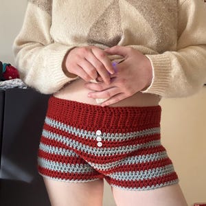 May include: Hand-crocheted shorts in a red and gray striped pattern. The shorts feature a high waist and three white buttons on the front. The garment is handmade and appears to be made of soft yarn.