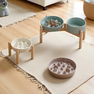 Peut inclure: Plusieurs gamelles pour animaux de compagnie de différentes couleurs et designs sont présentées sur un tapis beige clair. Une gamelle est bleu clair, une autre blanche et une troisième rose pâle. Les gamelles sont sur des supports en bois.