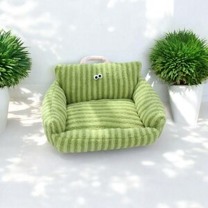 Peut inclure: Un lit pour animal de compagnie ray&eacute; vert avec une texture pelucheuse et une poign&eacute;e rose. Le lit a deux yeux noirs brod&eacute;s sur le devant.