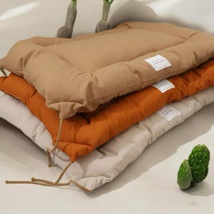 May include: Three quilted cushion pads in beige, orange, and white. The cushions are stacked on top of each other. The top cushion has a white label with black text.