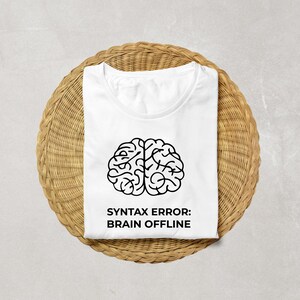 Peut inclure: T-shirt blanc avec un dessin de cerveau noir et le texte "SYNTAX ERROR: BRAIN OFFLINE". Le t-shirt pli&eacute; est pos&eacute; sur un support rond en osier brun clair. Le t-shirt a un col rond.