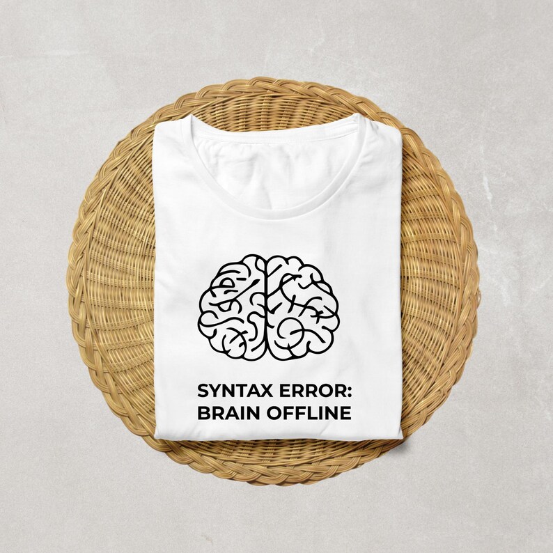 Peut inclure: T-shirt blanc avec un dessin de cerveau noir et le texte "SYNTAX ERROR: BRAIN OFFLINE". Le t-shirt pli&eacute; est pos&eacute; sur un support rond en osier brun clair. Le t-shirt a un col rond.