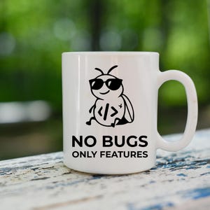Peut inclure: Mug en c&eacute;ramique blanche avec un dessin noir d'un insecte de dessin anim&eacute; portant des lunettes de soleil et tenant des symboles de code. Le mug porte l'inscription "NO BUGS ONLY FEATURES" en noir.