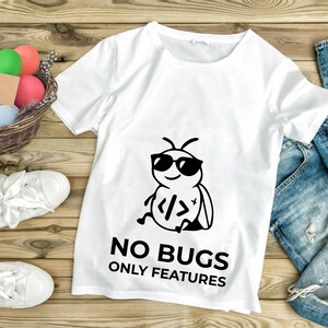 Peut inclure: T-shirt blanc avec un dessin noir d'un insecte portant des lunettes de soleil et des symboles de codage. Le texte "NO BUGS ONLY FEATURES" est imprim&eacute; en dessous. Le t-shirt est pos&eacute; sur une surface en bois.