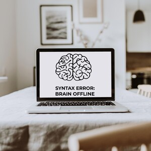 Peut inclure: Un ordinateur portable argent&eacute; ouvert sur une table. L'&eacute;cran affiche un cerveau noir et blanc avec le texte "SYNTAX ERROR: BRAIN OFFLINE". La table est recouverte d'une nappe claire.