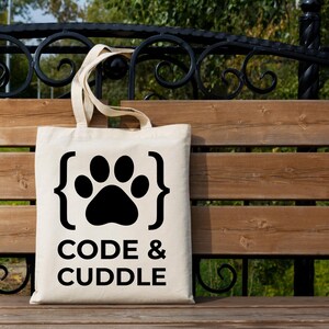Peut inclure: Un sac fourre-tout beige avec un motif de patte noire entre accolades et les mots "CODE & CUDDLE" imprim&eacute;s en dessous. Le sac a deux anses et est pos&eacute; sur un banc en bois.