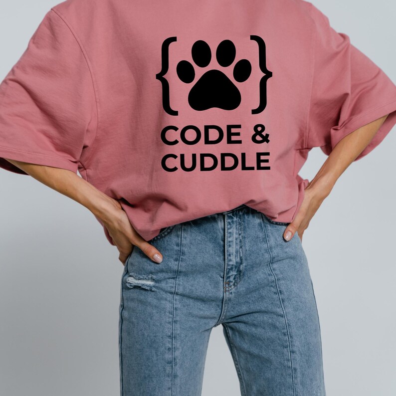 Peut inclure: T-shirt rose poudr&eacute; avec un motif de patte noire dans des accolades et l'inscription "CODE & CUDDLE" en gras. Le t-shirt est associ&eacute; &agrave; un jean en denim bleu.
