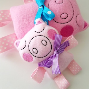May include: Two pink pig plush toys with embroidered details. The larger pig has a blue bow with white polka dots. The smaller pig has a purple bow with white stripes. Both pigs have pink and white polka dot ribbons.