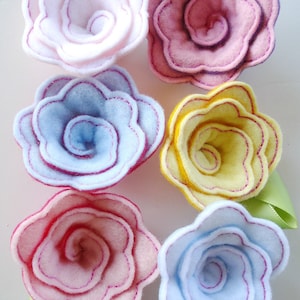 Pode incluir: Seis enfeites de flores de feltro em várias cores, incluindo rosa, azul, amarelo e branco. Cada flor tem um design em espiral em camadas com um ponto rosa ao redor das bordas.