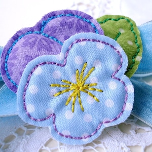 May include: Three fabric flowers in shades of purple, blue, and green. The blue flower has white polka dots and a yellow embroidered center.
