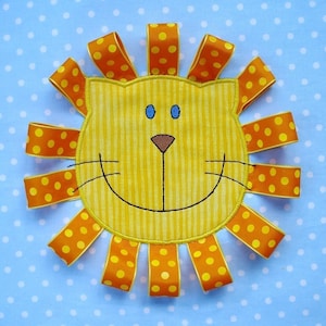 May include: A yellow felt lion head with a smiling face, blue eyes, and a black nose. The lion head is surrounded by yellow felt strips with orange polka dots.