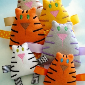 Puede incluir: Seis juguetes para gatos hechos a mano de fieltro en varios colores, incluyendo naranja, amarillo, blanco y gris. Cada gato tiene detalles bordados como rayas y ojos. Están adornados con cintas de colores.