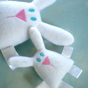 May include: Two white plush bunny toys with embroidered pink noses and blue eyes. The larger bunny has a white ribbon tied around its neck. The smaller bunny has white ribbon ties on its arms and legs.