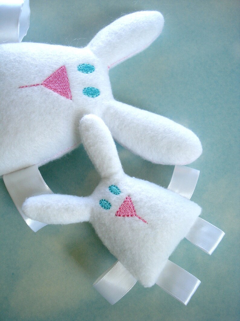 May include: Two white plush bunny toys with embroidered pink noses and blue eyes. The larger bunny has a white ribbon tied around its neck. The smaller bunny has white ribbon ties on its arms and legs.
