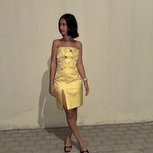 Puede incluir: Un vestido amarillo sin tirantes con un estampado floral y una abertura en la parte delantera. El vestido est&aacute; hecho de un material sedoso y tiene una silueta ajustada.