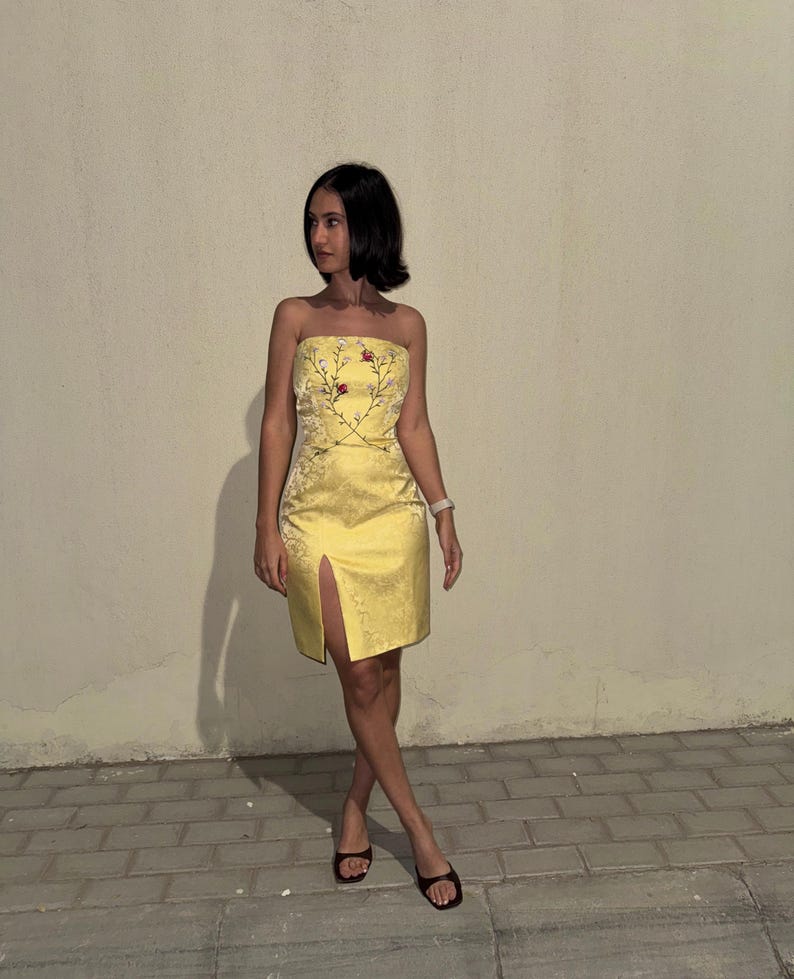 Puede incluir: Un vestido amarillo sin tirantes con un estampado floral y una abertura en la parte delantera. El vestido est&aacute; hecho de un material sedoso y tiene una silueta ajustada.