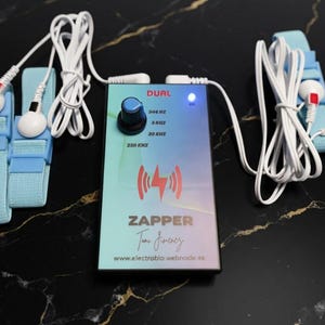 May include: A rectangular electronic device labelled DUAL ZAPPER with a blue control knob, a glowing blue indicator light, and text listing frequencies 344 HZ, 8 KHZ, 30 KHZ, 250 KHZ. It includes two white cables connected to light blue fabric straps.