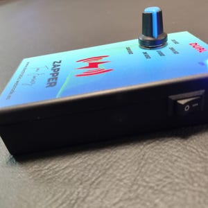 Puede incluir: Un dispositivo electrónico azul y verde con la palabra "ZAPPER" impresa en él. El dispositivo tiene una perilla, un interruptor y una luz LED azul. El dispositivo está etiquetado como "DUAL".