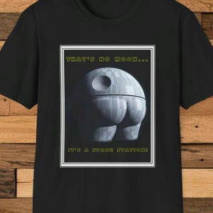Può includere: T-shirt nera con una grafica umoristica della Morte Nera e la scritta "THAT'S NO MOON... IT'S A SPACE STATION!" La grafica della Morte Nera è in un riquadro bianco.