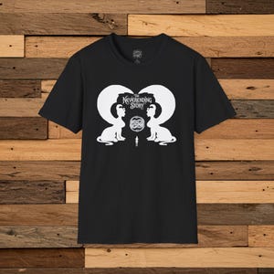 Może przedstawiać: Czarny t-shirt z białą grafiką z filmu "Niekończąca się opowieść". Projekt przedstawia dwie postacie podobne do sfinksów zwrócone twarzami do siebie, z małą postacią pomiędzy nimi. Tekst "The NeverEnding Story" znajduje się nad postaciami.