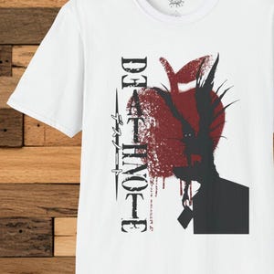 May include: White t-shirt featuring a graphic design with the text "DEATH NOTE" in black and a red apple with a silhouette of a character. The design has a grunge aesthetic.