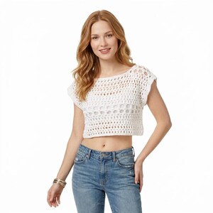 Könnte beinhalten: Ein weißes, kurzärmeliges, bauchfreies Häkeloberteil mit einem offenen Strickmuster und einem weiten, runden Ausschnitt. Das Kleidungsstück wird mit einer blauen Jeans kombiniert und wirkt durch die Textur handgefertigt.