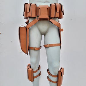 May include: A tan leather utility belt system featuring multiple rectangular pouches attached to the waist, thighs, and shins. The gear includes adjustable straps with brass-colored metal buckles, securing the various storage compartments to the legs.