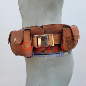 May include: A brown leather utility belt with two pouches and a gold-colored buckle. The belt is worn around the waist and is designed to hold various items. The belt is made of distressed leather.