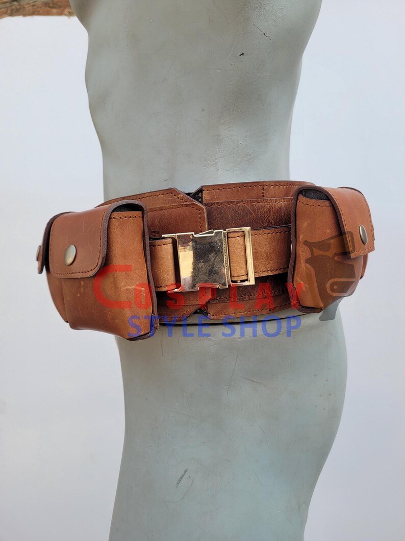 May include: A brown leather utility belt with two pouches and a gold-colored buckle. The belt is worn around the waist and is designed to hold various items. The belt is made of distressed leather.