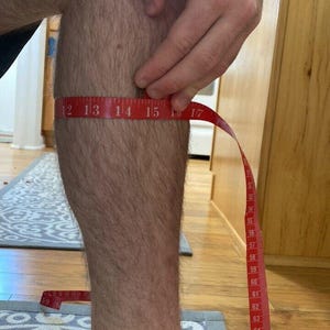 以下が含まれることがあります： 下腿部に巻き付けられた赤いメジャーは、周囲の長さが約39.5cmであることを示しています。メジャーには白い数字の目盛りが付いており、ふくらはぎ部分のサイズを示すために固定されています。