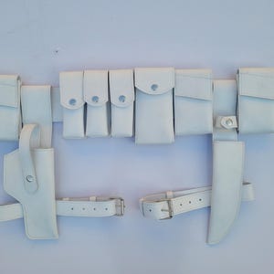 Peut inclure: Une ceinture utilitaire blanche dotée de plusieurs pochettes de tailles variées, d'un étui sur le côté gauche et d'un fourreau sur le côté droit. La ceinture comprend des sangles réglables avec des boucles métalliques et des boutons-pression.